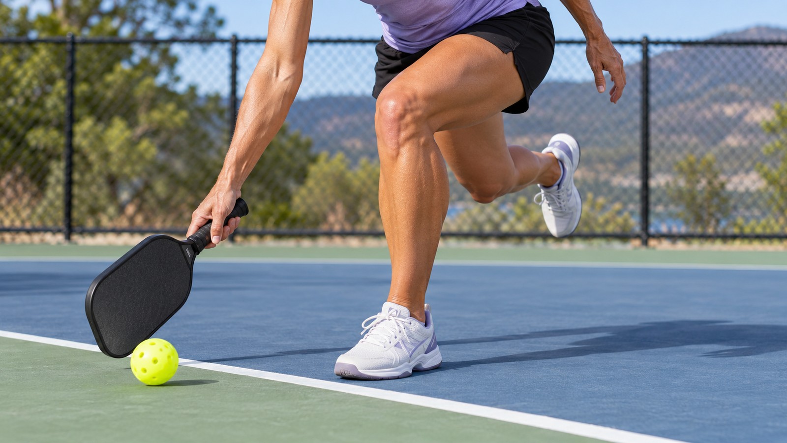 A person in a purple shirt and black shorts tracking a yellow pickleball with a paddle on an outdoor court.