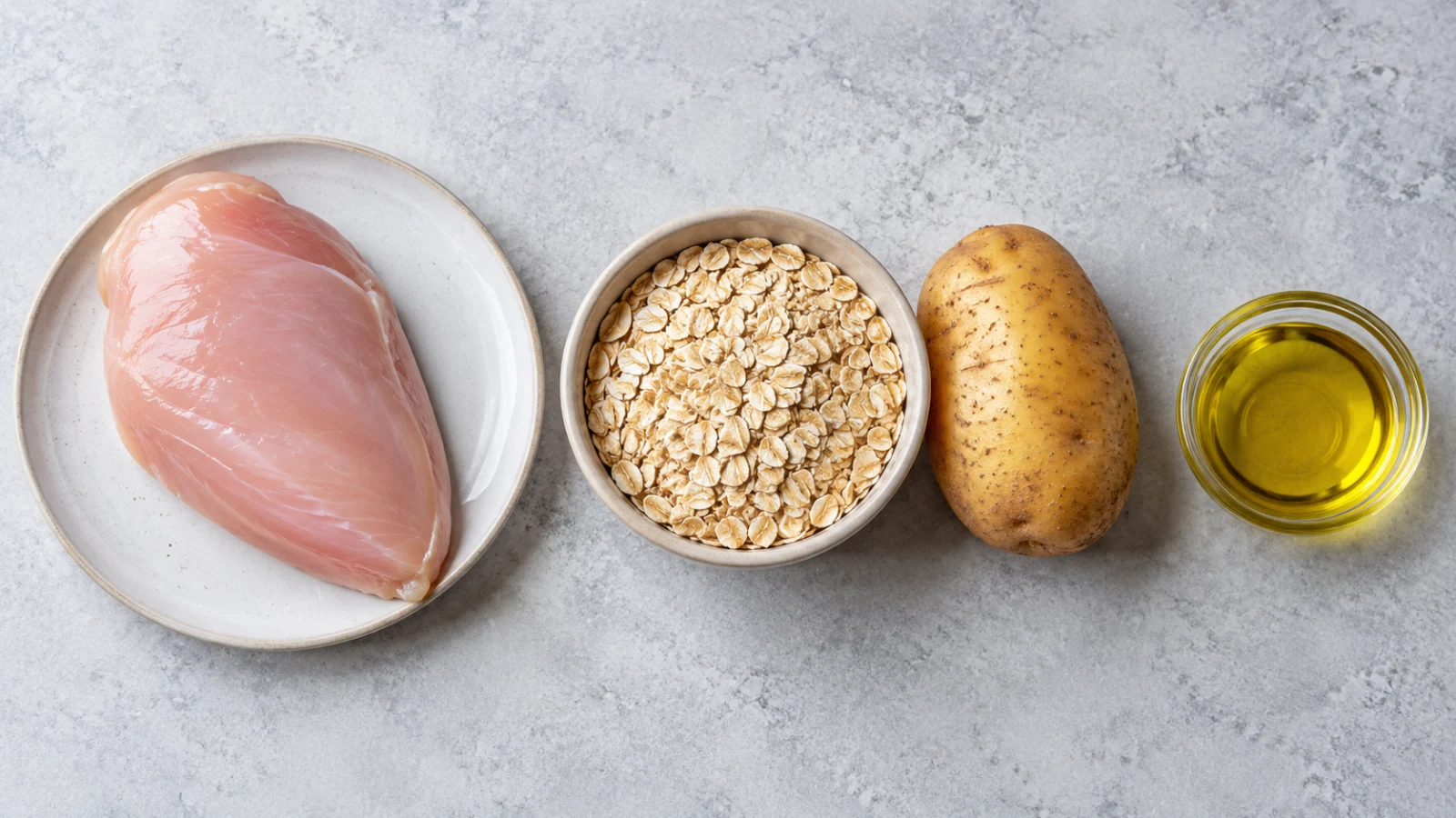 An overhead flat lay of a chicken breast, a bowl of oats, a whole potato, and a dish of olive oil.
